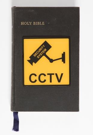 Image of Holy Bible with a blue ribbon hanging from the bottom and a yellow plastic CCTV sign adorned in the centre of the cover. 