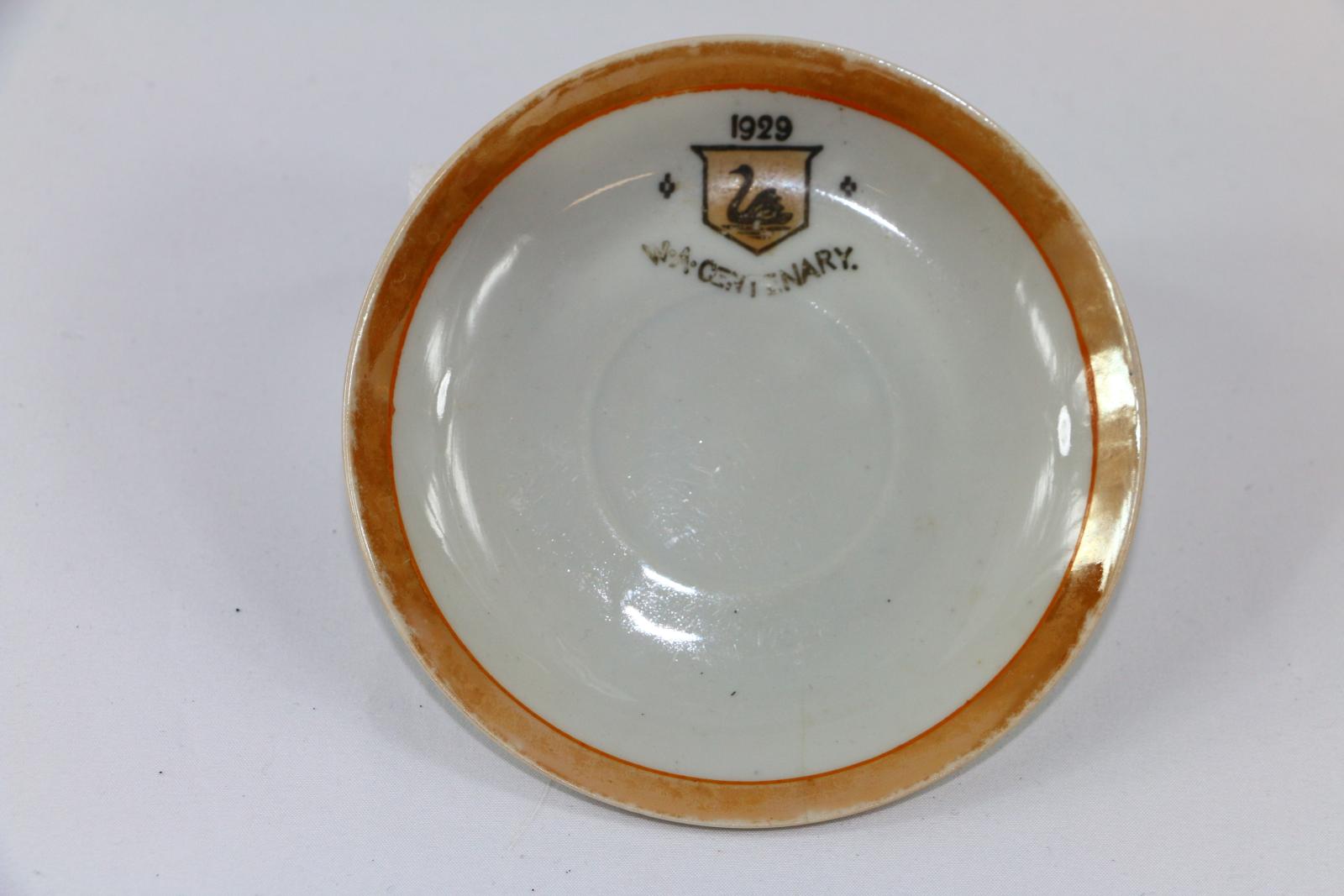 Small white, bone china saucer with glazed orange rim and emblem and shield on inside of saucer on one side.