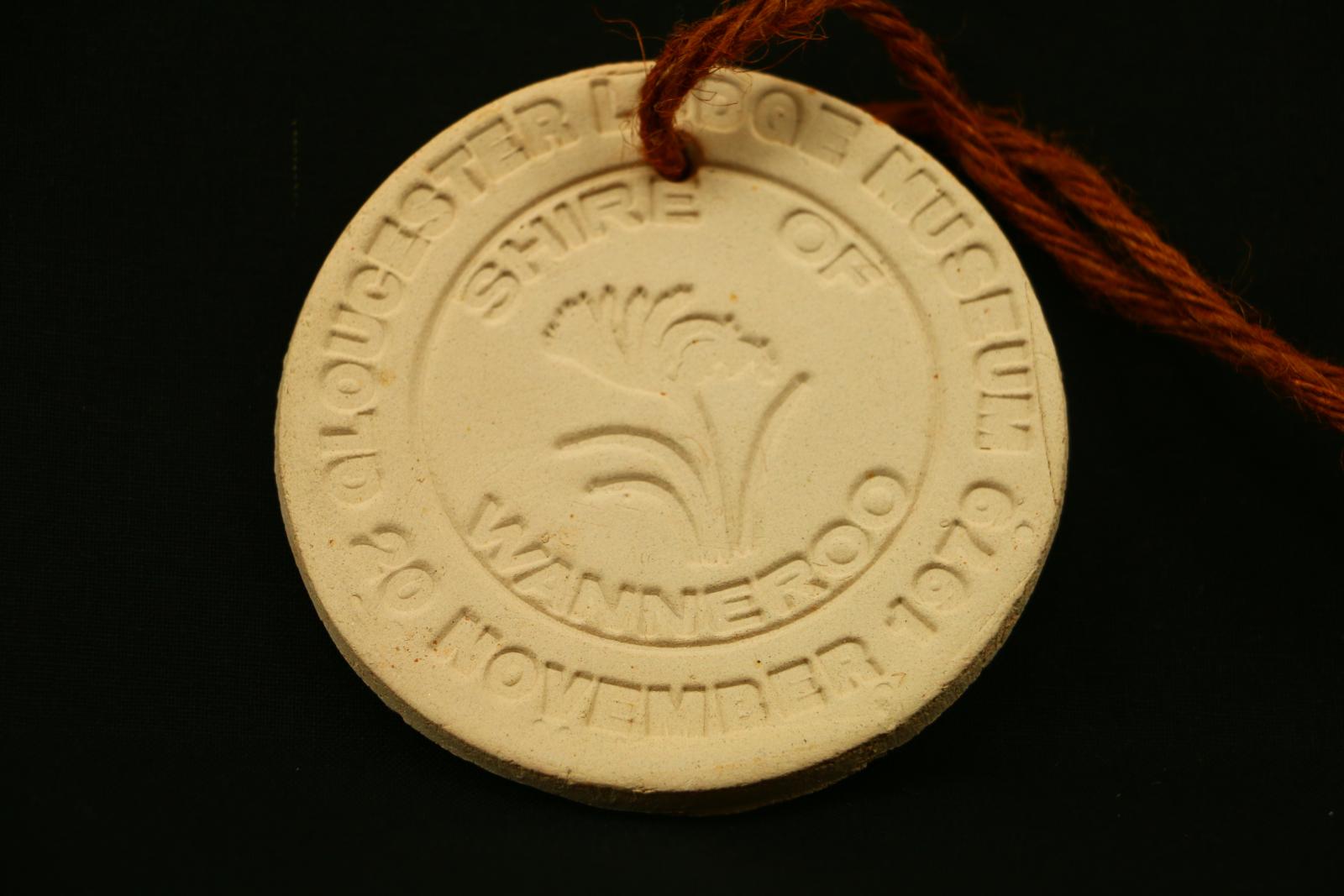 Small round clay pendant on a piece of brown string. It has an imprint in a circular pattern around the edge "GLOUCESTER LODGE MUSEUM 20 NOVEMBER 1979". Within an inner circle in a circular design "SHIRE OF WANNEROO" and the City of Wanneros's emblem, the Kangaroo Paw.