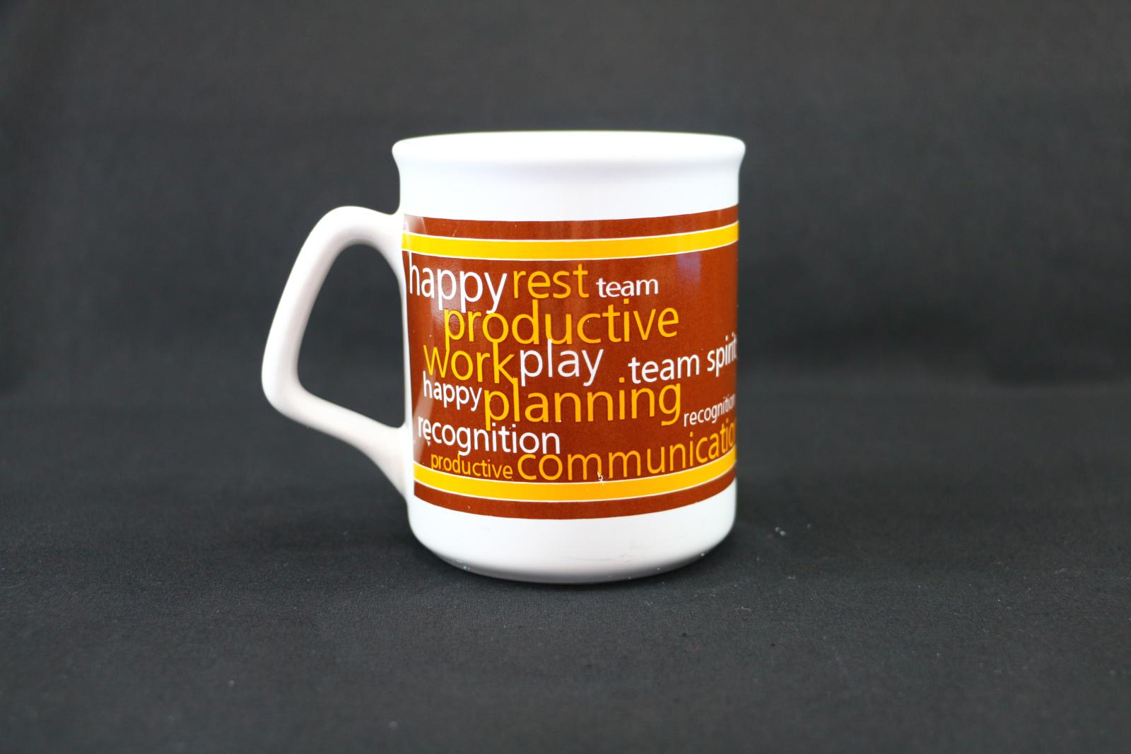 A white ceramic mug with a wide band of brown and yellow wrapped around the centre. The brown and yellow band has a selection of words printed in white or yellow, followed by the City of Wanneroo logo in white, and the staff conference logo, next to this is the year in which it was held in a yellow dot pattern with five thin white lines directly under the number, then in white with a brown nose is the design of a cow.