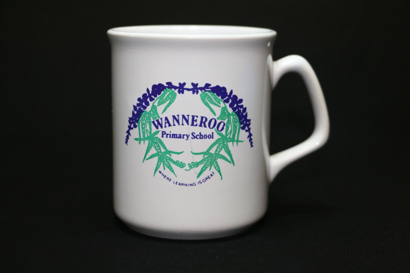 A white ceramic Wanneroo Primary School mug. One side of the mug has a round motif of purple flowers curving over the top with green leaves coming down from either side. In the middle in purple letters is 'WANNEROO/Primary School'. Then underneath the green leaves in very small purple letters 'WHERE LEARNING IS GREAT'