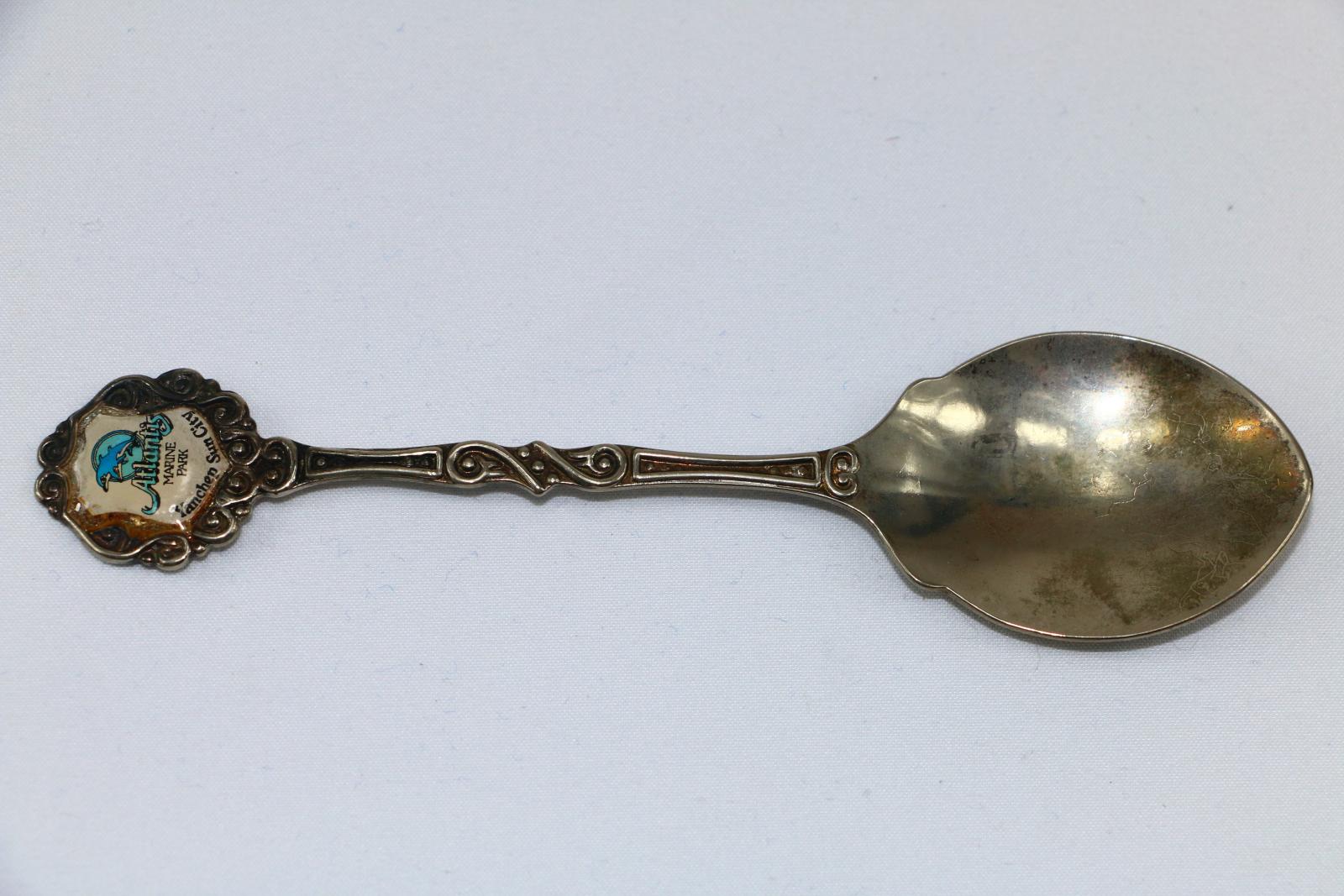 A long, thin, silver-plated souvenir teaspoon with an enamel badge on the handle. The bowl of the teaspoon is flatter with a small scalloped edge on each side which continues towards the front tip. The thin long-handle has raised edge line decorations of swirls and curved rectangles each side of the dots in the centre. The top of the handle is round with raised swirls and a white enamelled shield badge in the centre of 'ATLANTIS/MARINE/PARK'. On the lower edge of the badge is 'YANCHEP SUN CITY'. 