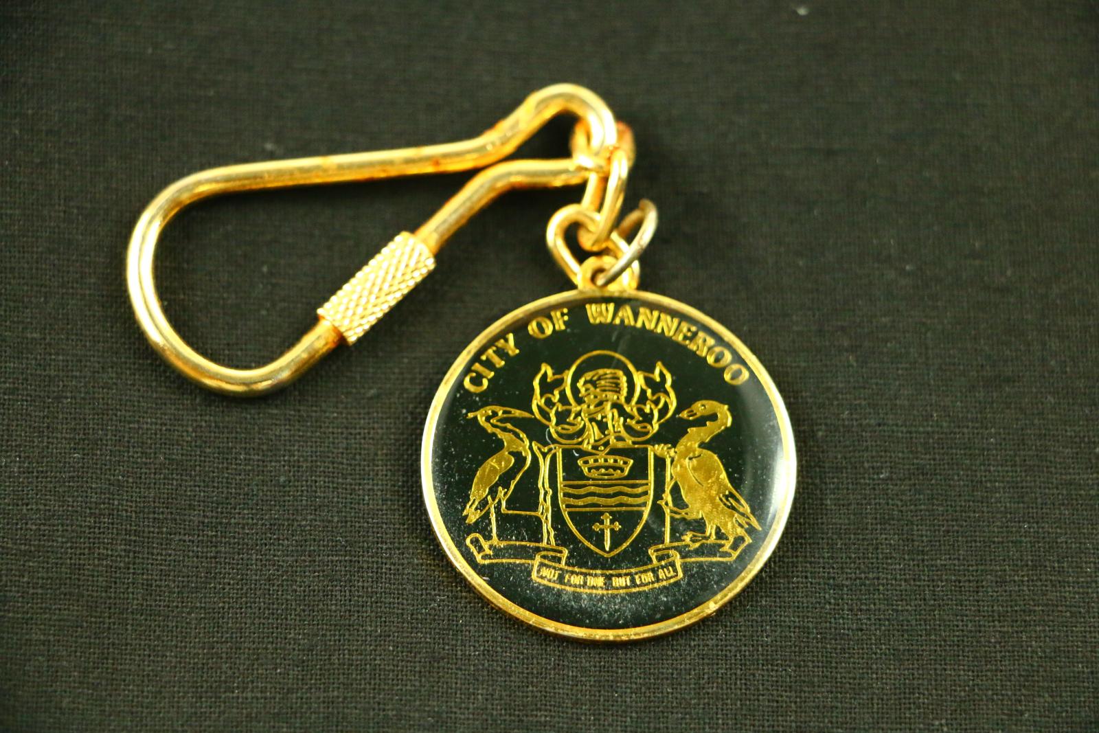 Round gold and black, metal, City of Wanneroo key ring. Gold coloured, carabiner attached to one end with a small screw clasp. On the other end of the chain is a round, gold metal disc. The front of the disc is enamelled in black with a thick clear lacquer coating. In the centre of the disc is a crest with two tall water birds holding a shield between them in gold. On the shield 'CITY OF WANNEROO' and 'NOT FOR ONE BUT FOR ALL' is written.. 