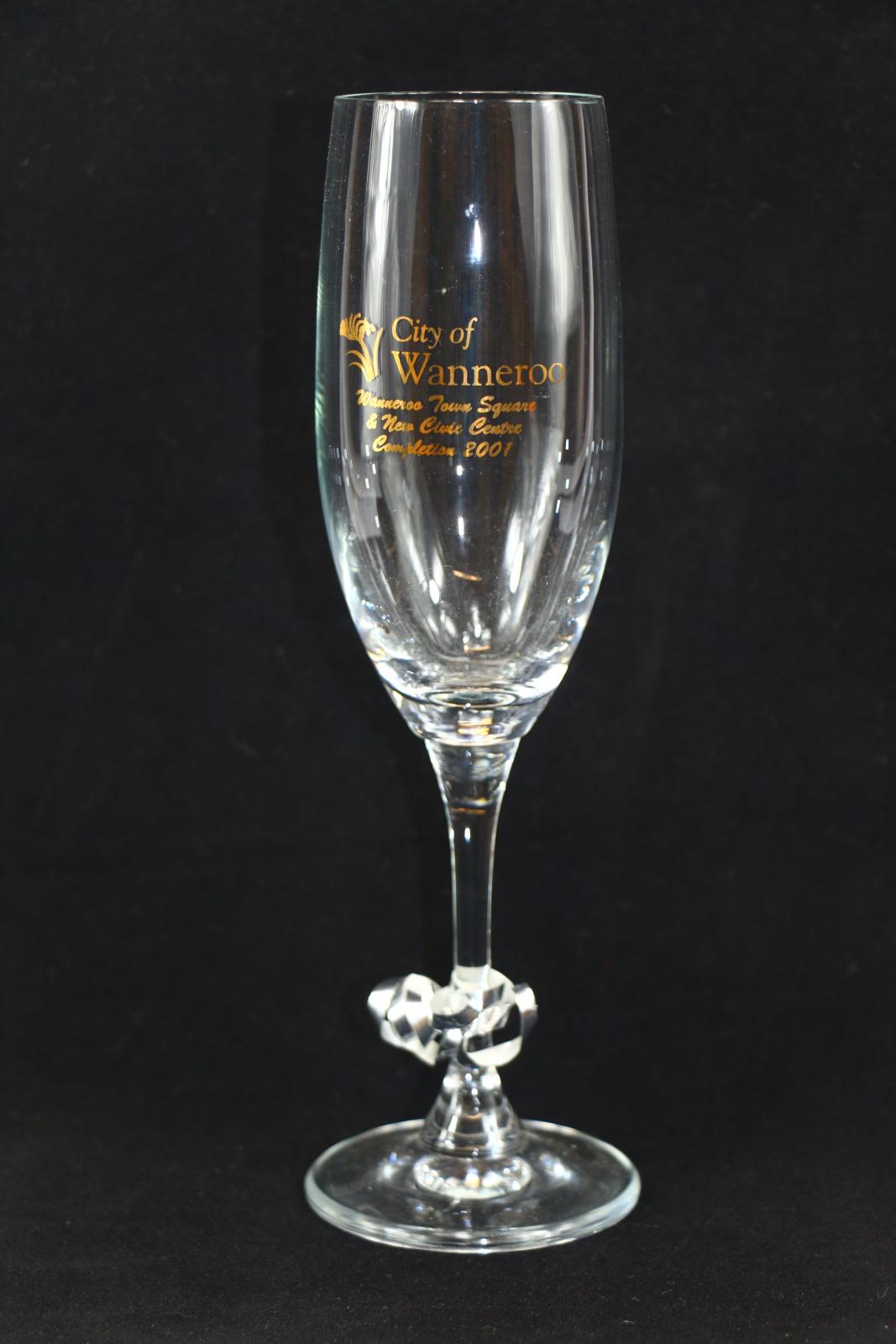 A City of Wanneroo champagne flute. Printed on the front of the glass in gold is a kangaroo paw flower motif. To the right of the motif in gold lettering is 'City of Wanneroo'. Underneath this and the motif in cursive gold script is 'Wanneroo Town Square & New Civic Centre Completion 2001'. Tied around the stem of the glass is a short piece of silver coloured curling ribbon, curled tightly at each end.