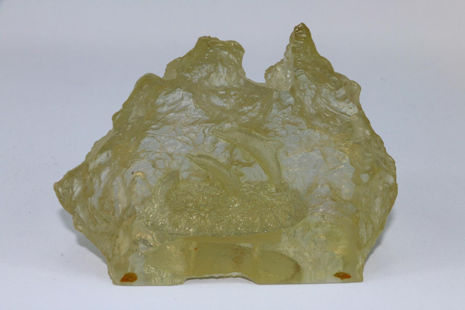 A moulded glass object in the shape of Australia with a smooth cut cross-section showing two dolphins leaping from water suspended within moulding. The object has a flat base with three very small felt pieces glued to underneath.