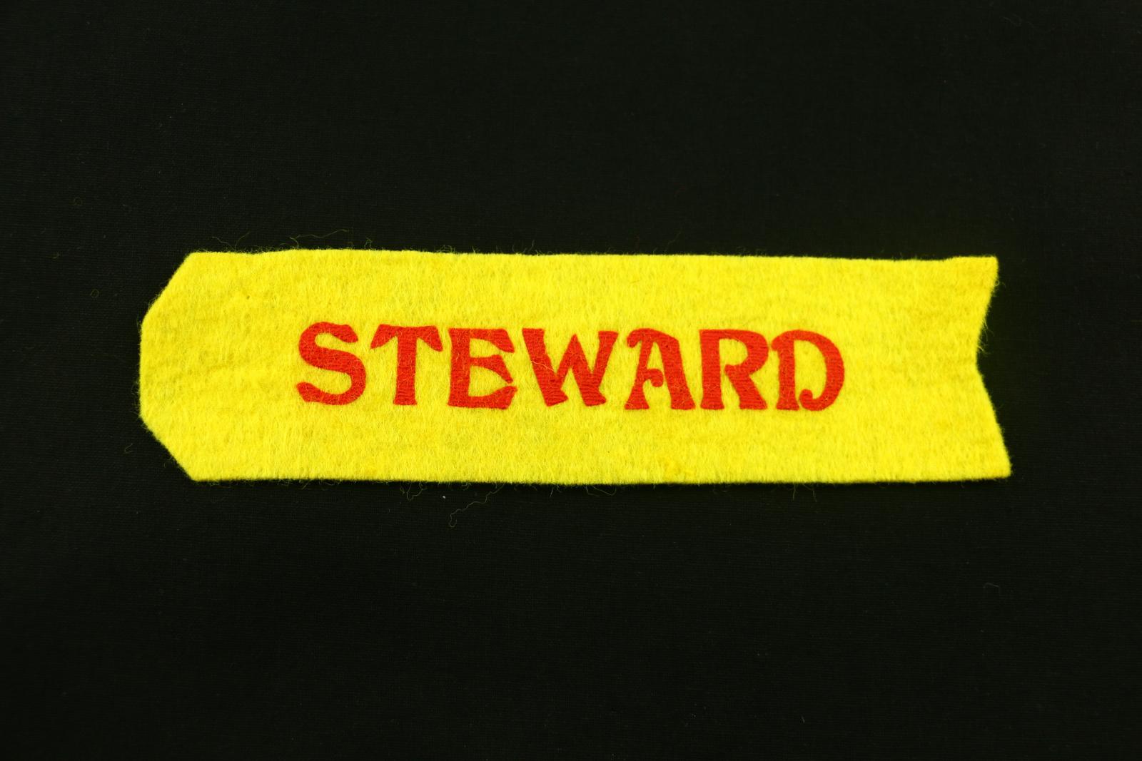 A yellow felt 'ribbon' with the word 'STEWARD' printed in red on one side of the object.