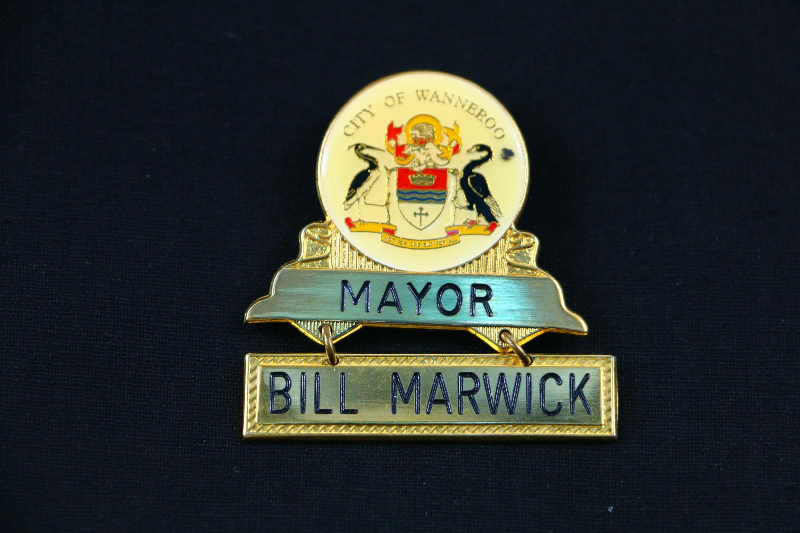 A gold metal badge with crest of City of Wanneroo. Underneath the crest the word 'MAYOR' IS ENGRAVED. Below the word MAYOR attached by two circular loops is a metal bar with the words 'BILL MARWICK', engraved on metal.