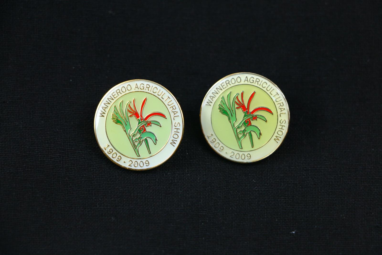 Gold metal pin with enamelled green and red kangaroo paw on front and 'WANNEROO AGRICULTURAL SHOW 1909-2009' written in gold around the edge of the pin, there is a butterfly clasp which attaches to the reverse of the pin.