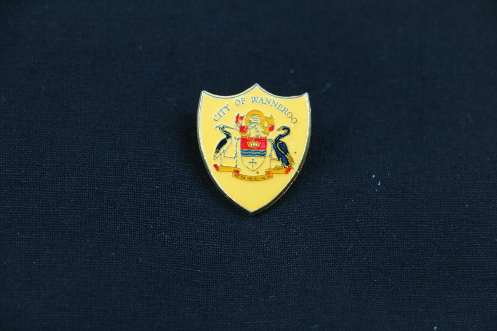 A  shield, with red, black and yellow coloured enamelled crest and butterfly clip on a lapel pin. The background of the three point shield is yellow with the words 'CITY OF WANNEROO' across the top. The crest of Wanneroo covers the main foreground showing two birds, a shield, and a scrolled ribbon. The yellow and red scrolled ribbon has the words stamped in black ink, 'NOT FOR ONE BUT FOR ALL'. 