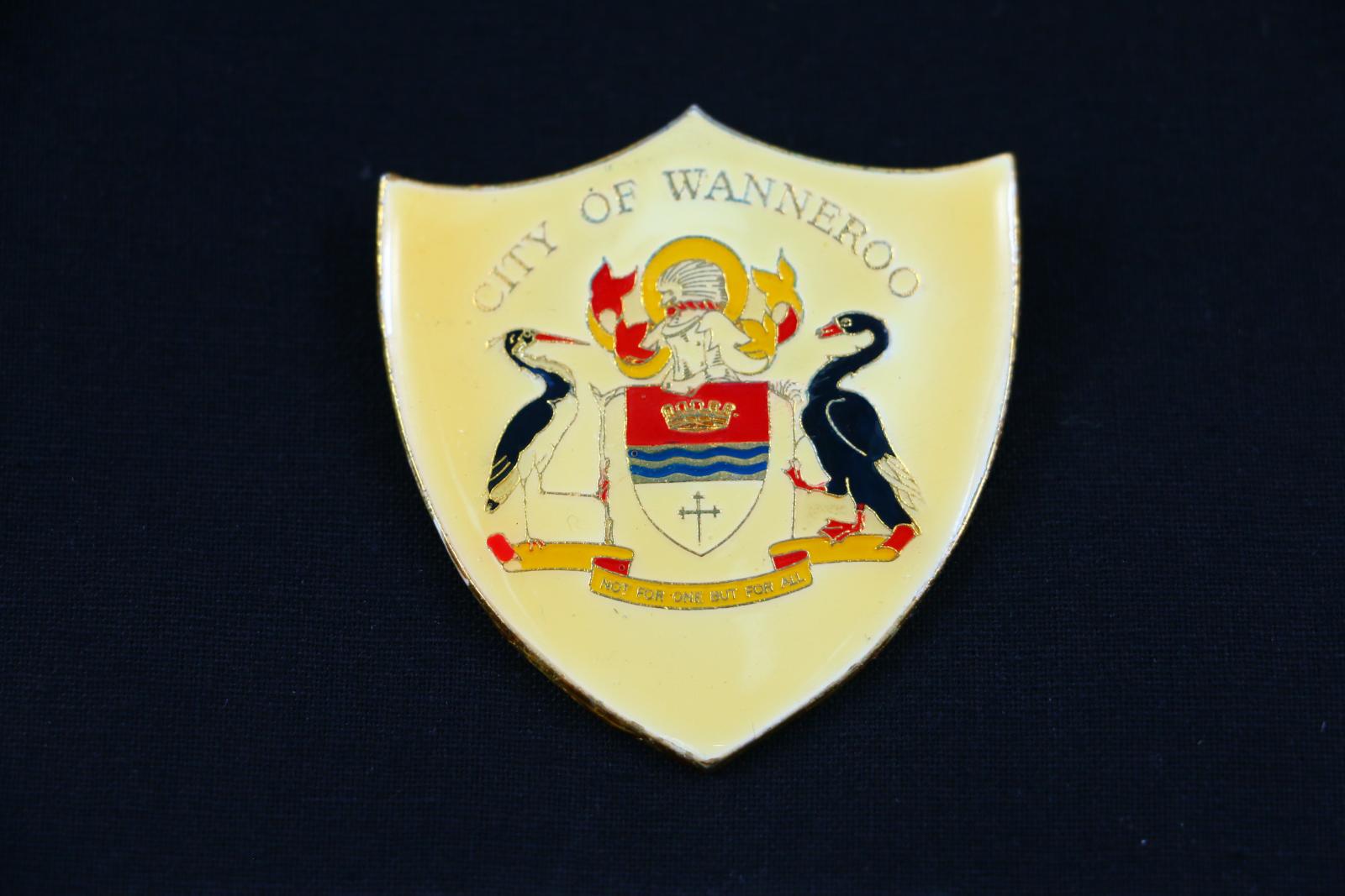 A shield, with red, black and yellow coloured enamelled crest and butterfly clip on a lapel pin. The background of the three point shield is yellow with the words 'CITY OF WANNEROO' across the top. The crest of Wanneroo covers the main foreground showing two birds, a shield, and a scrolled ribbon. The yellow and red scrolled ribbon has the words stamped in black ink, 'NOT FOR ONE BUT FOR ALL'.