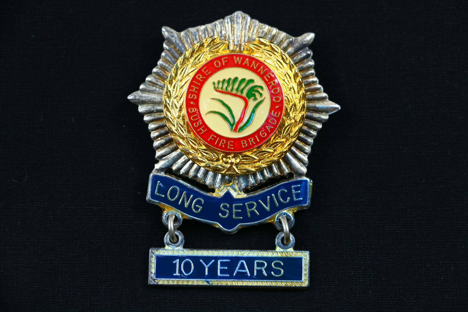 A silver, enamel, eight point badge with an attached bar. The silver outer part of the badge consists of eight scalloped sections with six graduating pressed points in between each scallop. The inner section of the badge has a red enamel outer edge with 'SHIRE OF WANNEROO BUSH FIRE BRIGADE' written in gold and a green and red kangaroo paw in the centre. The bottom of the badge has a bar which states 'LONG SERVICE' and attached by metal links underneath is a badge that states '10 YEARS' both written in gold 