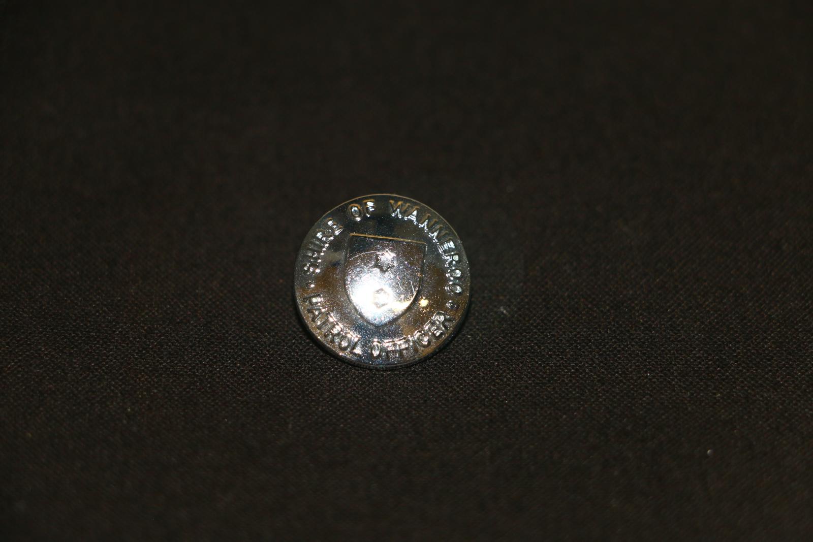 Silver metal, round button with the words, 'SHIRE OF WANNEROO' embossed around the top edge and 'PATROL OFFICER' around the lower edge. There is a raised shield in the centre which has two stars embedded and evenly placed. 