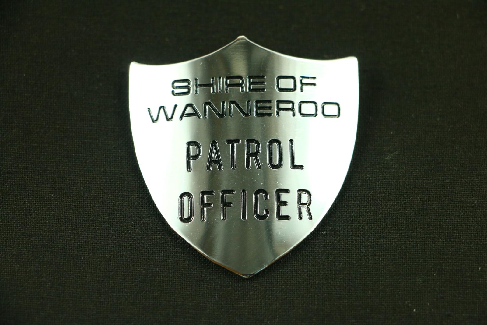 A silver metal three point shield badge. The badge has the words 'SHIRE OF WANNEROO PATROL OFFICER' etched in black ink on the front.