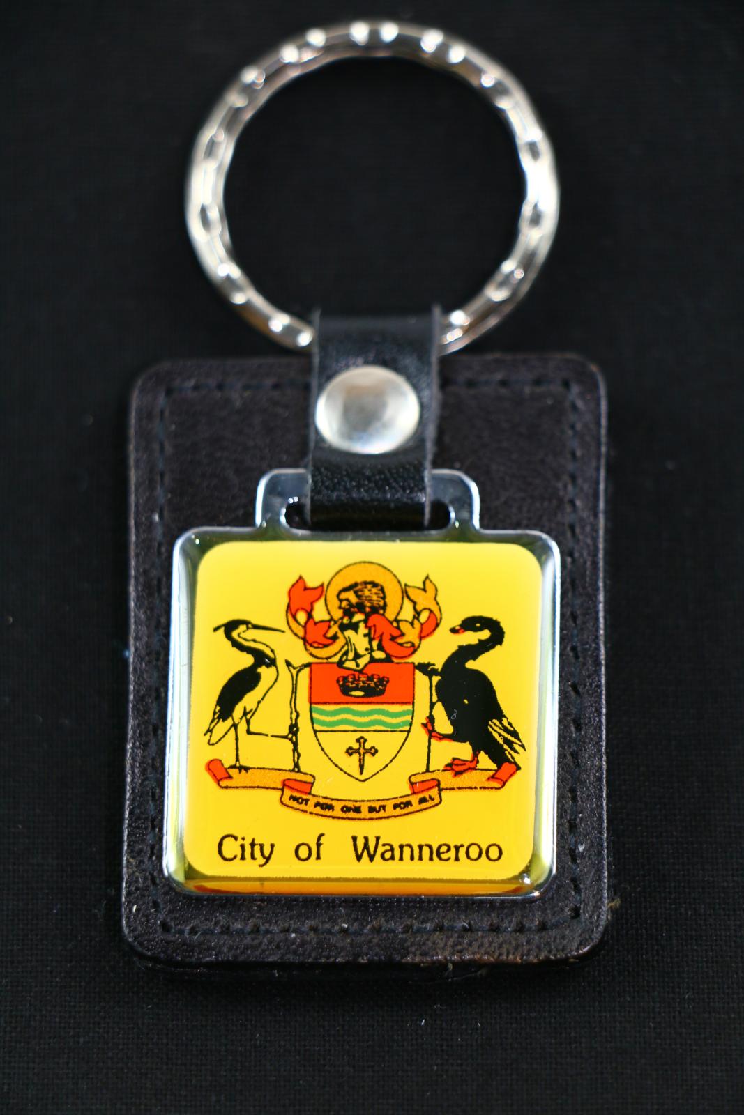 A City of Wanneroo keychain, which has a black leather rectangle backing with a square silver metal attachment on the front which has the City of Wanneroo crest. The crest consists of two birds facing each other with a shield in the centre, there is a scroll at the bottom of the birds that says 'NOT FOR ONE BUT FOR ALL'  and underneath the crest 'City of Wanneroo' is written.