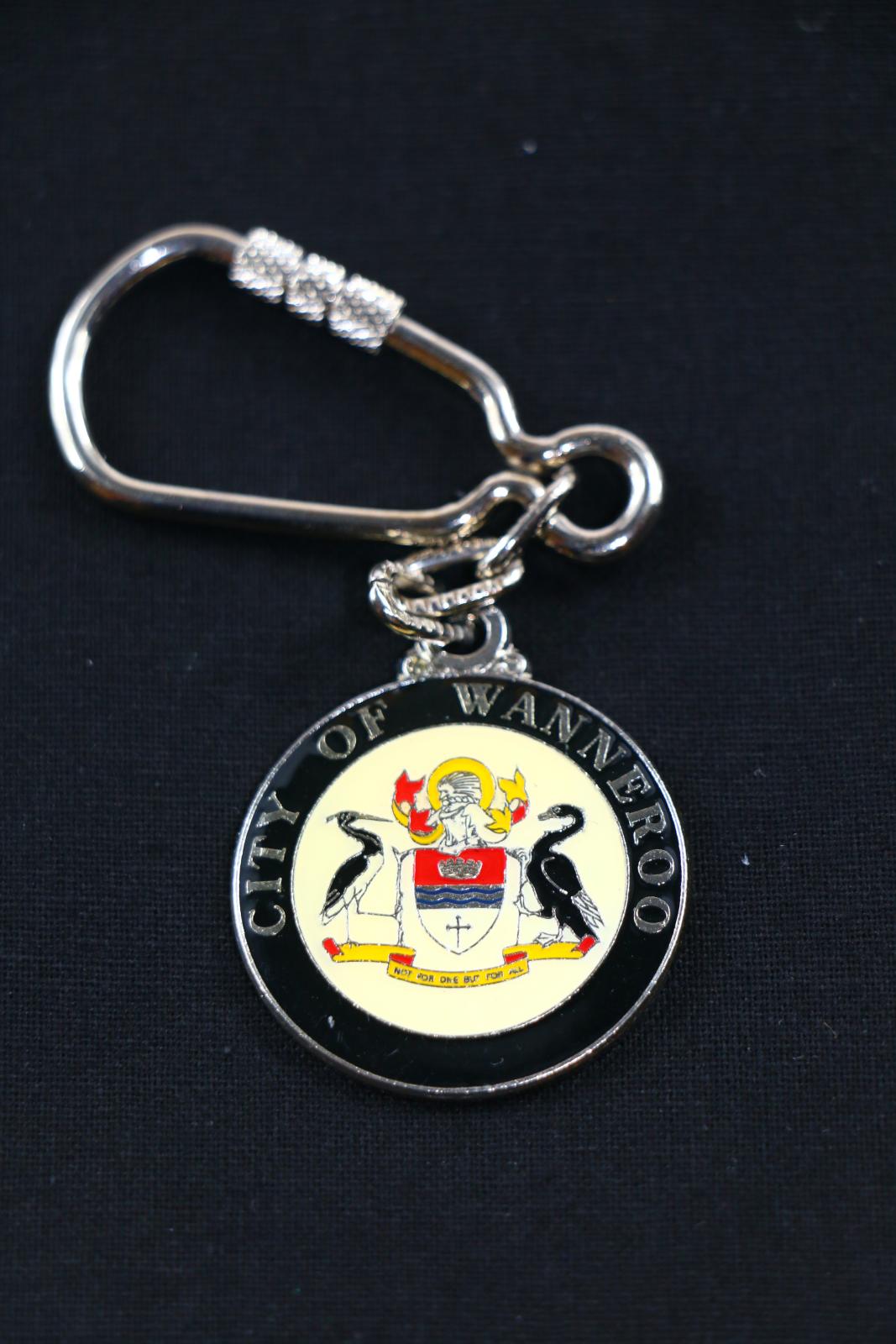 A silver metal City of Wanneroo medallion keychain attached to a silver chain and carabineer. The border of the medallion is black with 'CITY OF WANNEROO' written in silver. Within the border is the City of Wanneroo crest on a beige background. The crest consists of two birds facing each other with a shield in the centre, there is a scroll at the bottom of the birds that says 'NOT FOR ONE BUT FOR ALL'.