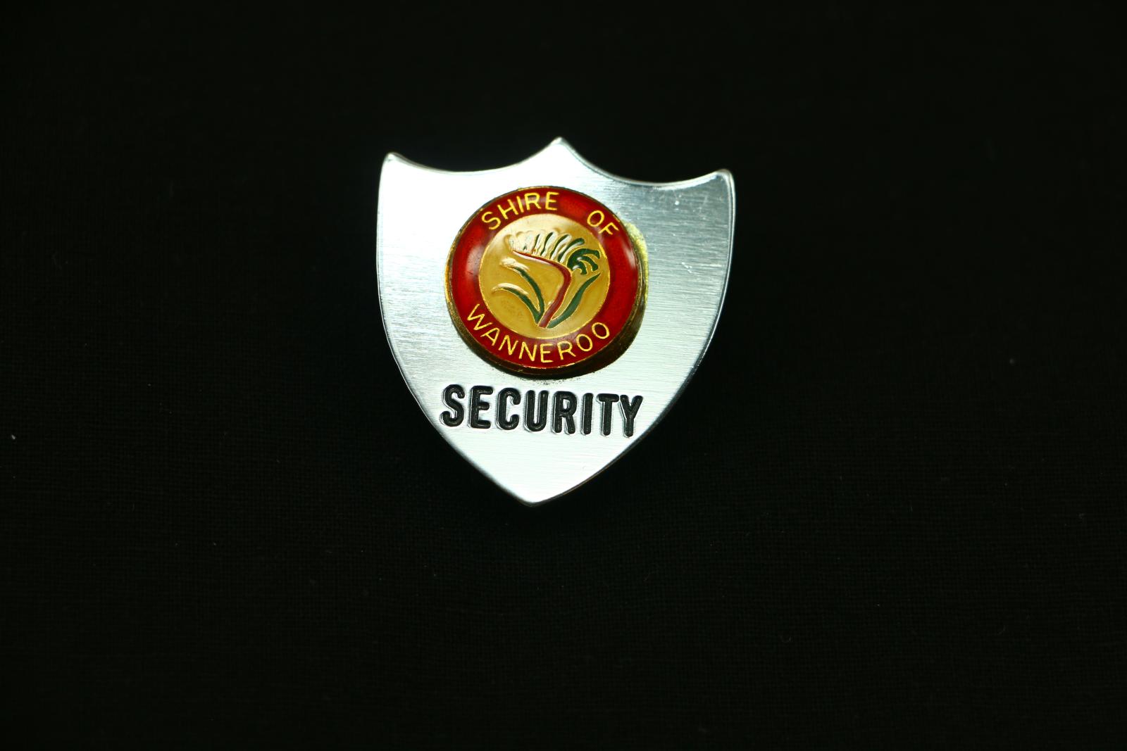 A silver metal three point shield badge. The badge has a circular enamel badge in the centre which has a red border and 'SHIRE OF WANNEROO' written in gold within, the centre has a red and green kangaroo paw. The bottom of the badge has 'SECURITY' imprinted and painted with black ink.  