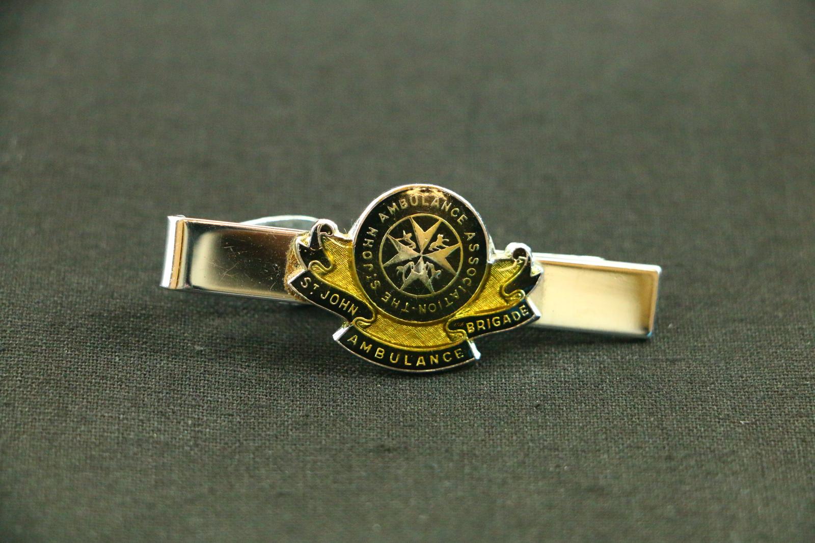 A silver metal St John Ambulance tie clip. Long, thin, silver metal bar with a seated clip attached to the back from a hinge on the left side. On the front of the bar is an small, round enamel badge, it has a dark blue circle in the middle with a silver outline and a stylized cross in the centre. Around the edge of the circle in small silver letters is 'ST JOHN AMBULANCE ASSOCIATION'. Underneath is a long stylized ribbon with a dark blue background and a small silver letters, 'ST JOHN AMBULANCE BRIGADE'.