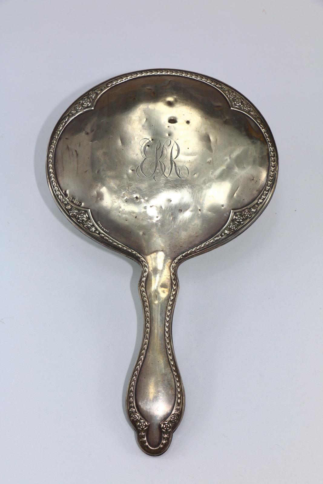 The back of a silver mirror with decorative edge and K.R engraved in the centre.