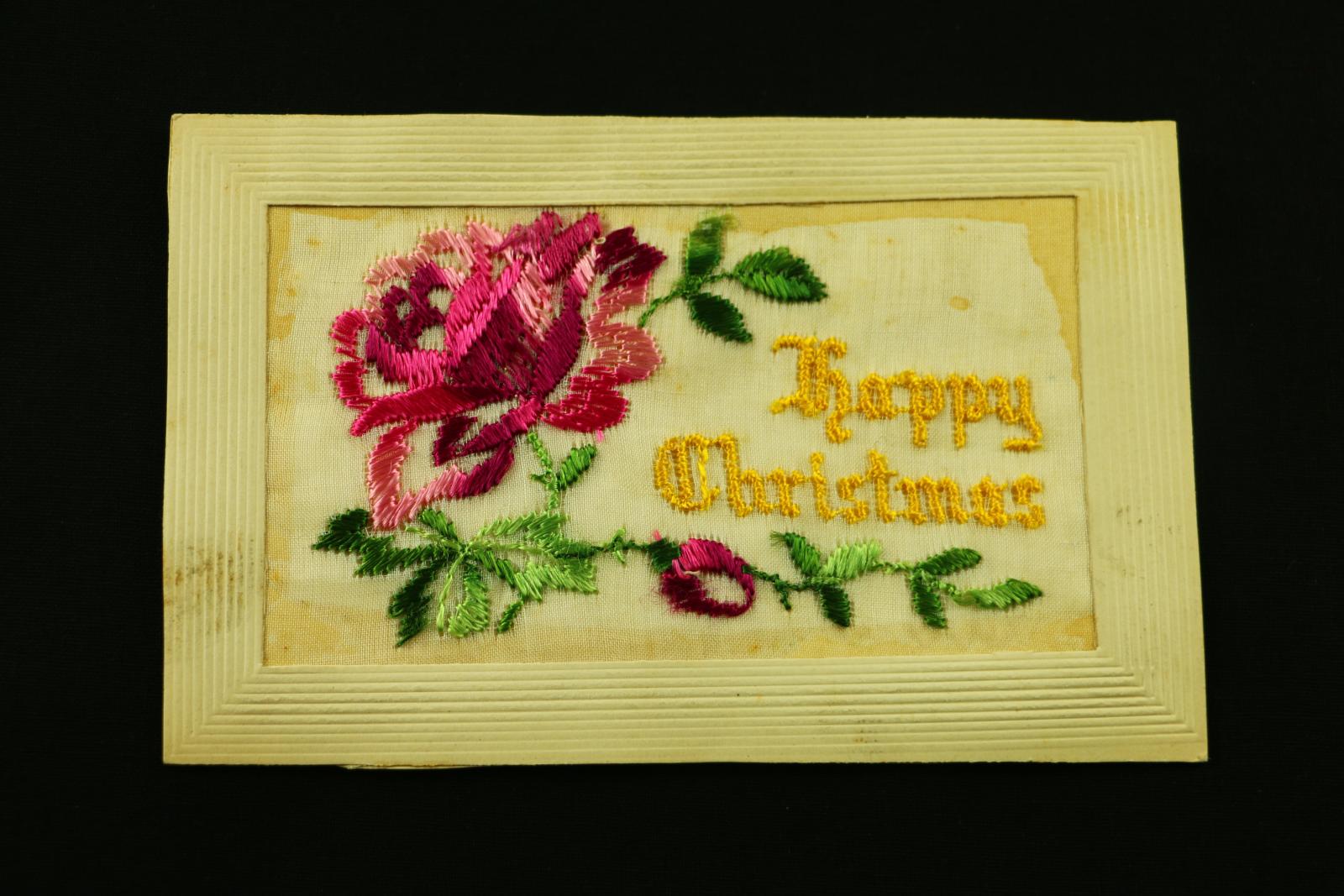 Rectangular, cardboard and embroidered 'Happy Christmas' postcard. The front of the postcard has a pink and red flower with green leaves embroidered on the front held within a cardboard frame, on the right is 'Happy Christmas' embroidered in yellow.