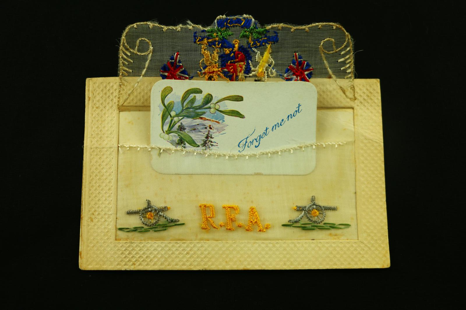 A rectangular, cardboard and embroidered, postcard R.F.A. UK crest and two small cannons on bottom corners. Inside the embroidered envelop is a small rectangular white card displaying a small branch of blossom with the words 'Forget me not' written in blue ink.