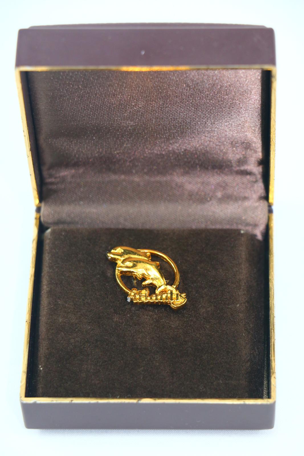 A circular, gold metal, Atlantis Marine Park souvenir pin in a brown box. The front face of the pin consists of a thin, gold, circular border with two leaping dolphins in the centre, the bottom part of the pin, has the text 'Atlantis', in cursive script, moulded in gold metal. The leftmost tail of the 'A' is broken off. A pin protrudes from the reverse side of the dolphin figures.