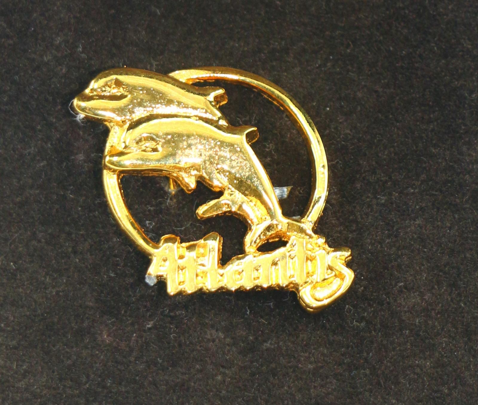 A close up image of the dolphin pin, which shows two gold dolphins in a circle and the word Atlantis along the bottom.