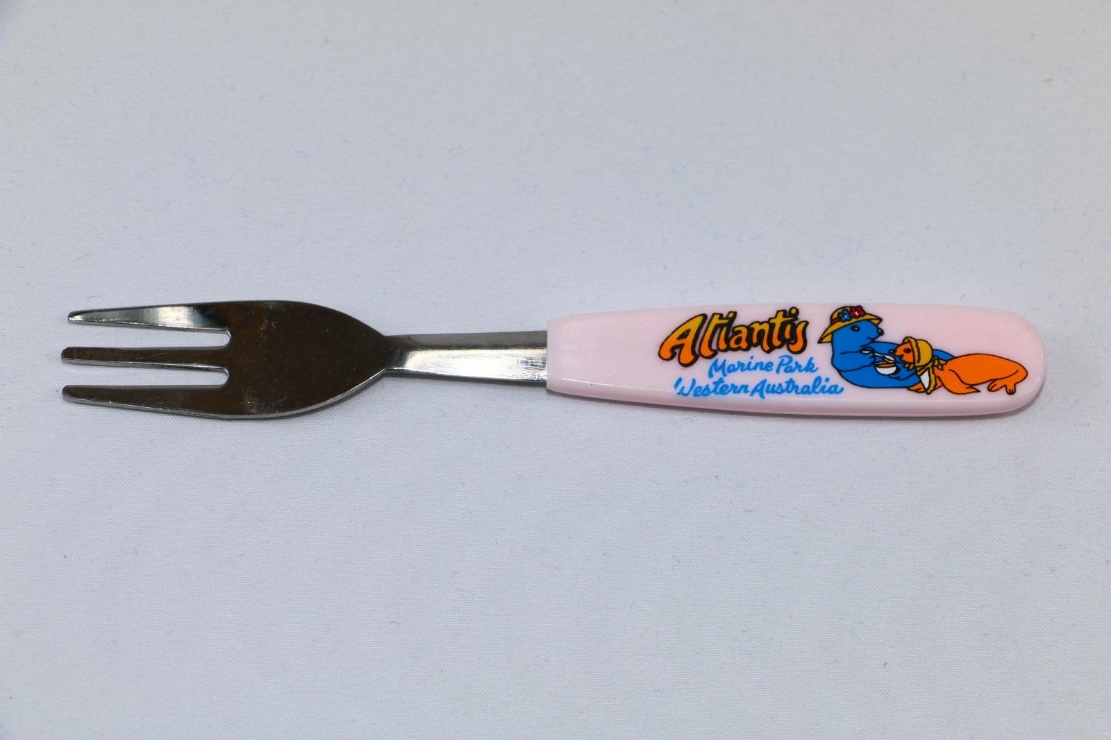 One small fork with a pink plastic handle, there is an image of two seals wearing hats, one blue mother seal feeding her orange baby seal from a bowl with a spoon and the words 'Atlantis Marine Park Western Australia'