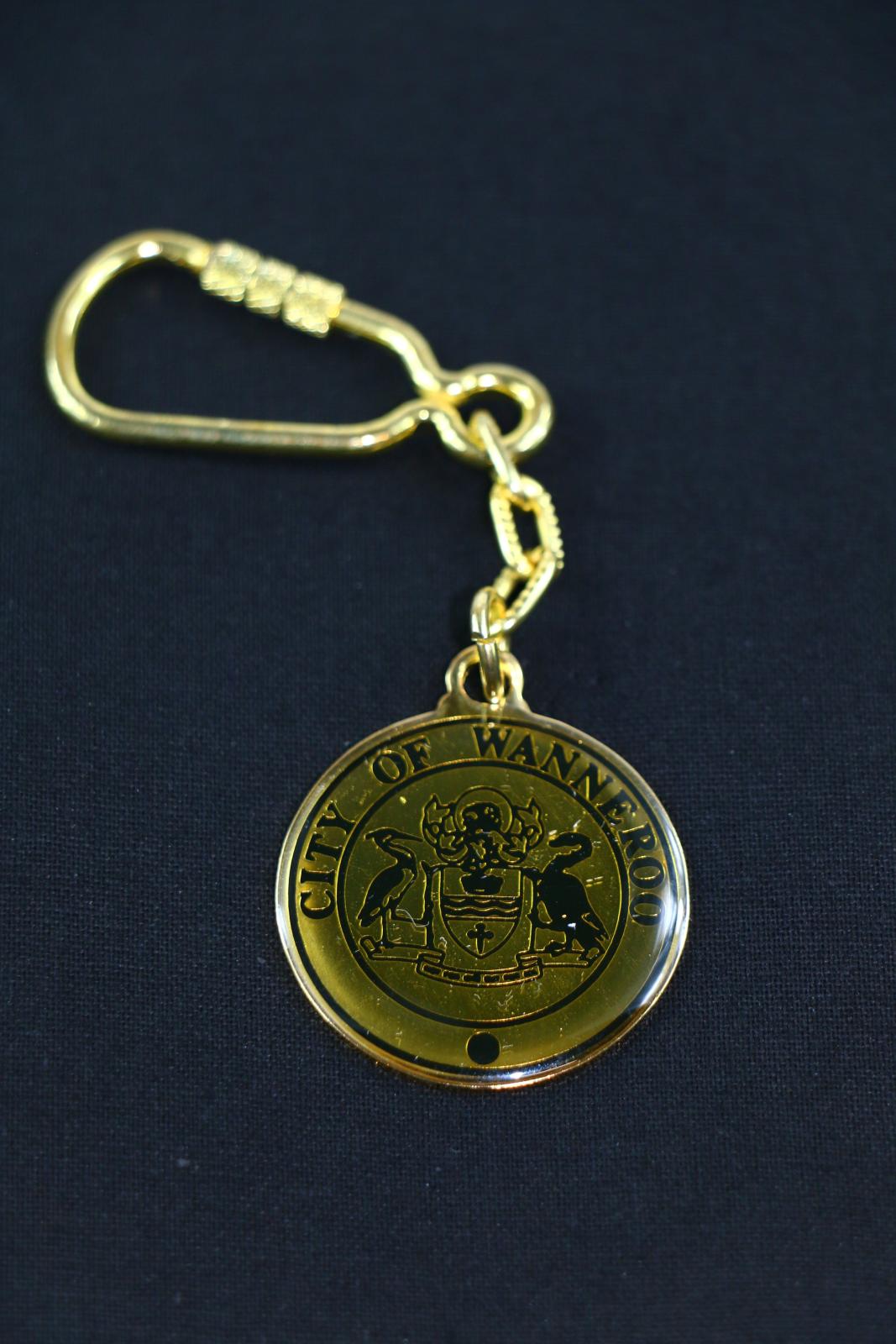 A gold metal City of Wanneroo medallion keychain attached to a gold chain and carabineer. The border of the medallion has 'CITY OF WANNEROO' written in black. Within the border is the City of Wanneroo crest. The crest consists of two birds facing each other with a shield in the centre.