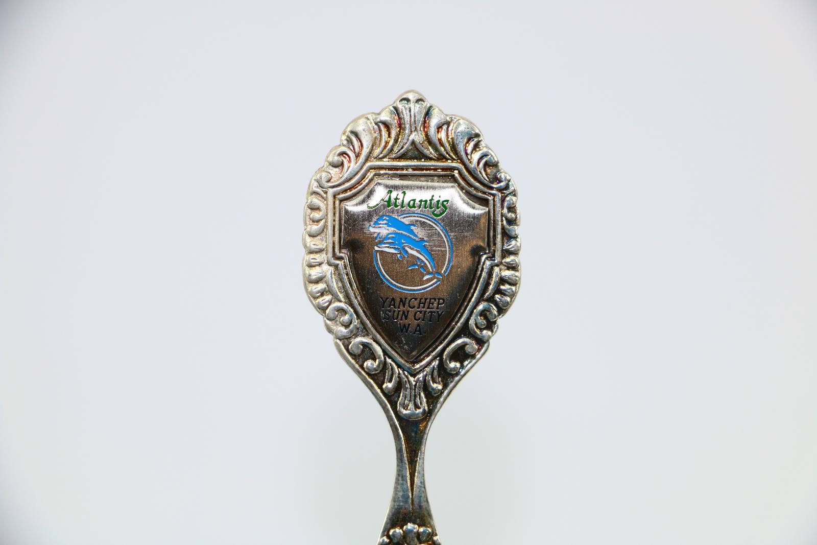 A close up image of the enamel badge at the top of the spoon. There is a blue and white image of two dolphins jumping in front of a circle. It also has the text 'Atlantis' in blue lettering at the top. Underneath the image of the dolphins is the text 'YANCHEP/SUN CITY/W.A'