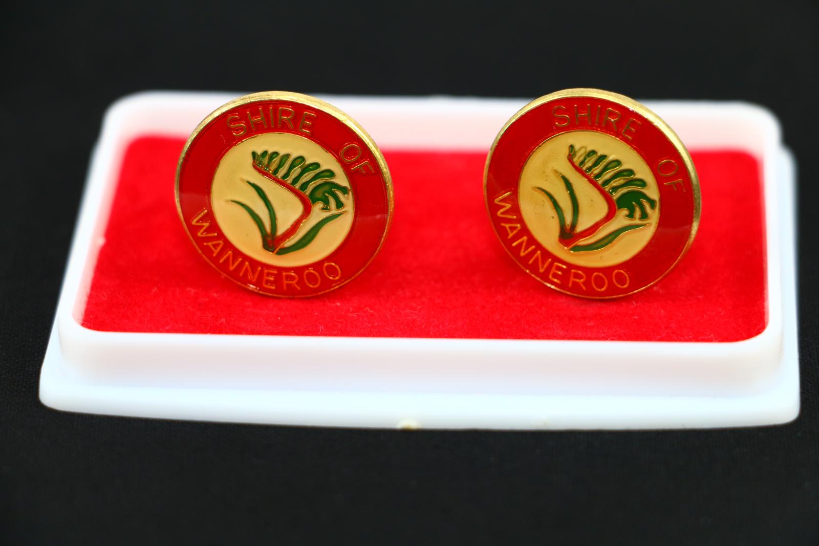 Gold-coloured metal cufflink with short toggle and post. The cufflinks have an enamel face which shows an image of a green and red kangaroo paw (the Shire emblem) with gold outlines on a light yellow circular background. Surrounding this image is a thick red border with the text 'SHIRE OF' on the top half, and 'WANNEROO' on the bottom half. The cufflinks sit in a plastic case, the lid is clear and the base is white and lined with red felt, there are two split plastic posts which hold the cufflinks in place.