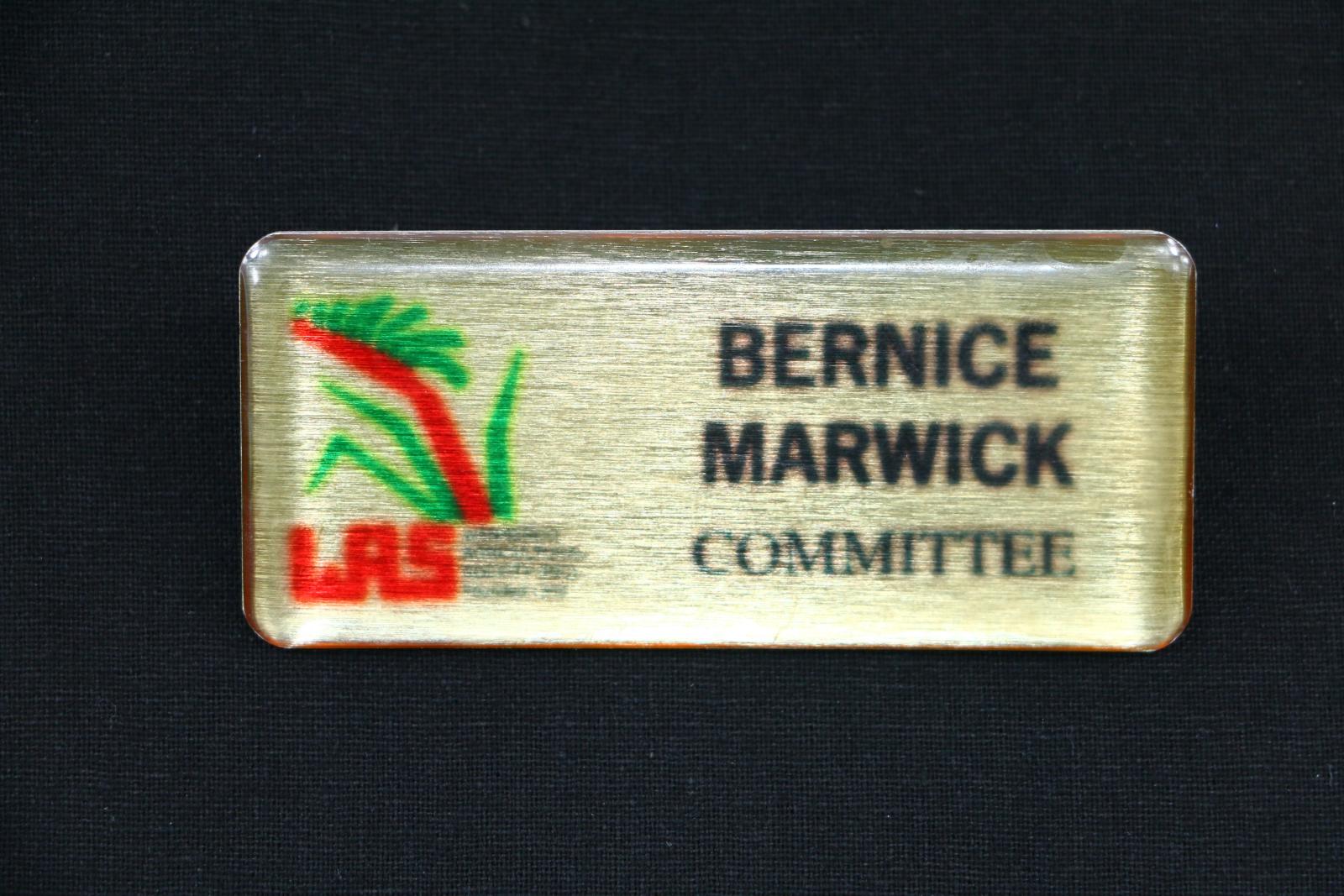 Rectangular, gold coloured plastic name badge with rounded corners. The face of the badge is shiny faux 'brushed metal' overlaid with thick clear plastic. The left hand side of the badge face is a red and green kangaroo paw symbol. Underneath this symbol is a red stylised acronym 'WAS' and very small black text which reads 'WANNEROO AGRICULTURAL SOCIETY (INC) Founded 1909'. On the right hand side of the badge face is large bolded black text reading 'BERNICE MARWICK' and underneath reads 'COMMITTE'.