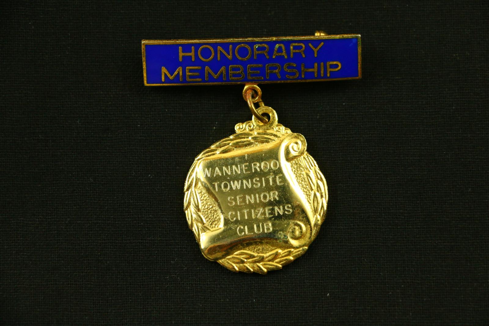 A gold metal badge which has a long, thin, horizontal rectangle inset with blue enamel and gold lettering, under which a gold coloured metal circular medal hangs from one circular metal chain link. The front of the rectangle reads 'HONORARY MEMBERSHIP'. The front of the circular medal has a relief of a scroll against a laurel wreath background. On the scroll is engraved 'WANNEROO TOWNSITE SENIOR CITIZENS CLUB'. The back of the circular medal is engraved 'Mrs. BERNICE MARWICK'.