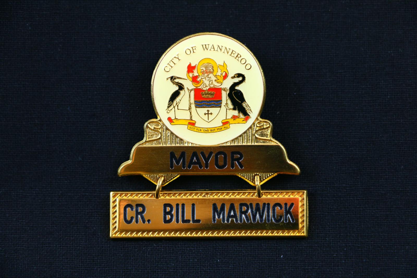 Gold coloured metal badge with a cream coloured enamel circle bearing the City of Wanneroo coat of arms, above a loose rectangular name plate attached by two golden metal chain links. The top of the circle reads 'CITY OF WANNEROO' in small gold lettering above the City's coat of arms. Text on the badge reads 'MAYOR' and 'CR. BILL MARWICK'.