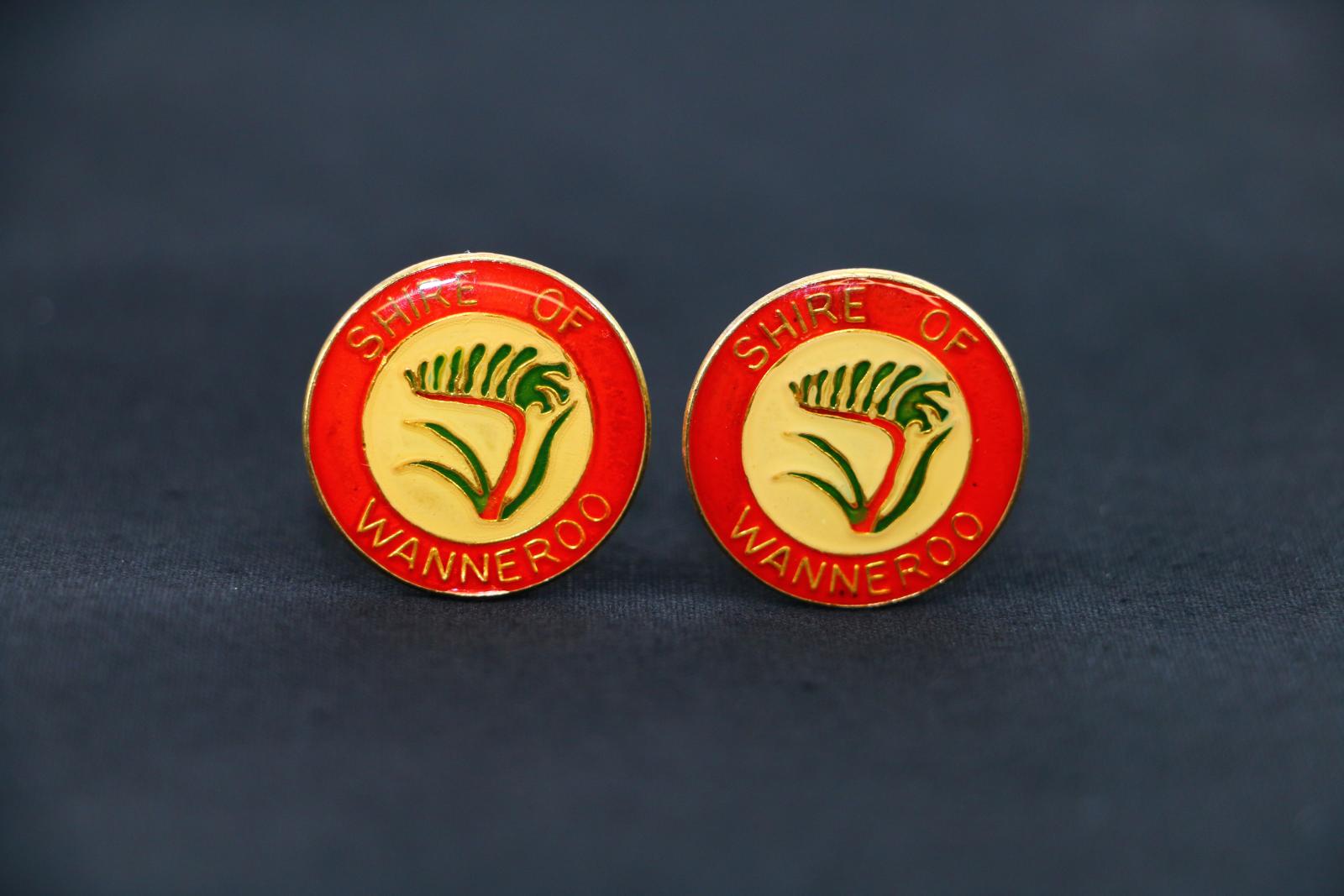 Gold coloured metal cufflink with a post attached to a hinged, moveable toggle on the back, and coloured, circular enamel front face. Enamel face shows central image of green and red kangaroo paw (the Shire emblem) with gold outlines on a light yellow circular background. Surrounding this image is a thick red border with the text 'SHIRE OF' on the top half, and 'WANNEROO' on the bottom half. 