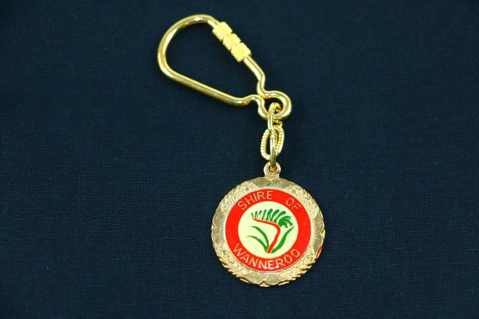 Gold coloured metal keyring with round disc hanging from a short chain off a large clasp. The face of the disc has a gold coloured metal border with four shallow decorative relief sections resembling foliage spaced evenly around it. This border surrounds a central circular enamel panel. The face of this panel shows a central image of a green and red kangaroo paw (the Shire emblem) with gold outlines on a light yellow background. Surrounding this image is a thick red enamel border with the text 'SHIRE OF' on