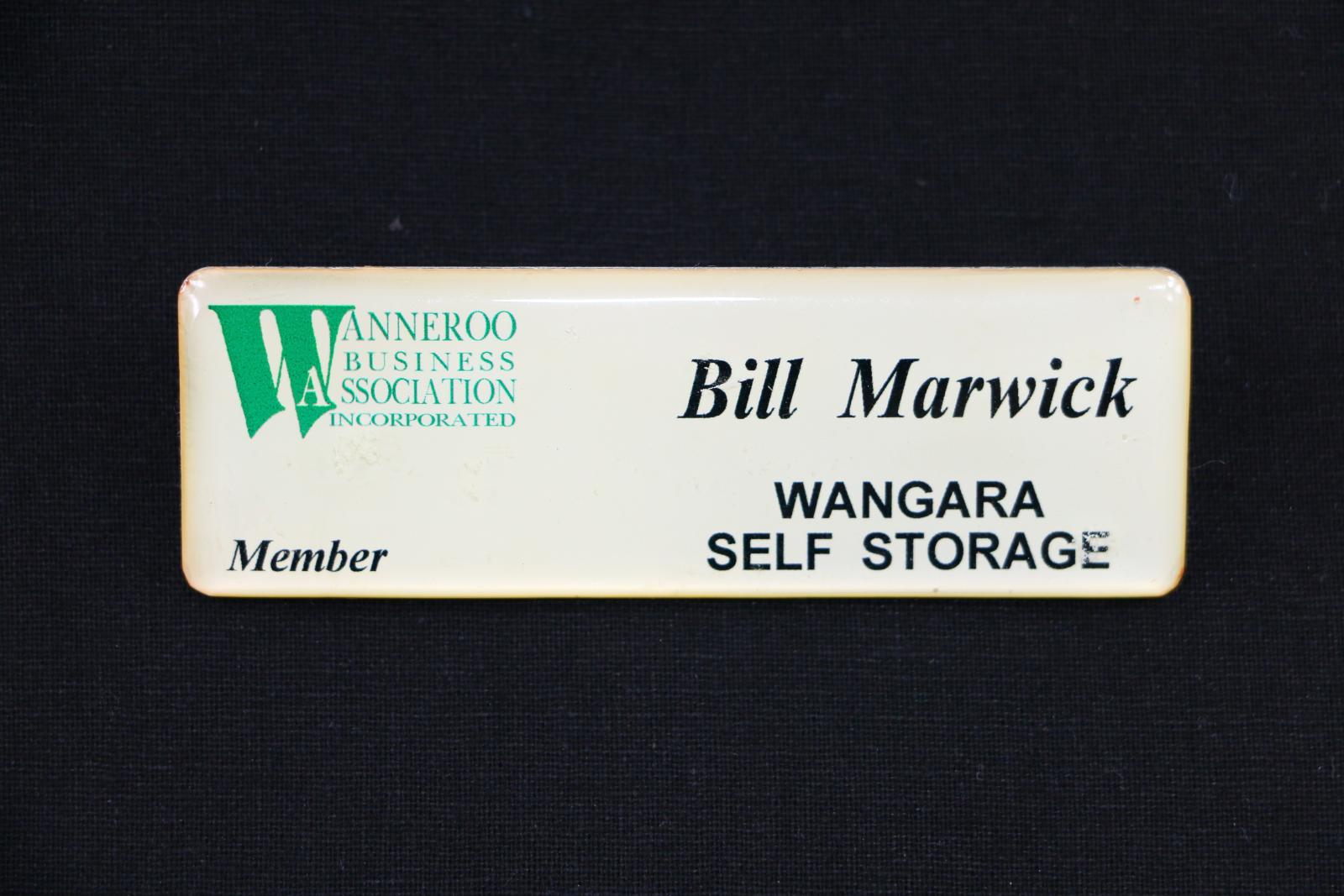 Rectangular, cream coloured plastic name badge with rounded corners. The front of the badge is pearlescent cream overlaid with thick clear plastic. On the upper left hand corner of the badge is a green logo which has small text next to a large 'W'. The logo text reads 'WANNEROO BUSINESS ASSOCIATION INCORPORATED'. On the bottom left hand corner is italicised black text which reads 'Member'. On the upper right hand corner is italicised black text which reads 'Bill Marwick'. 