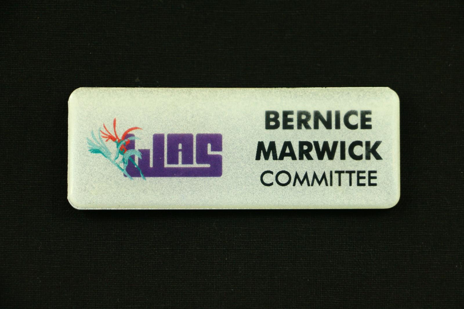 Rectangular, silver coloured plastic name badge with rounded corners. Face of badge is glittery silver overlaid with thick clear plastic. On the left hand side is a logo featuring an purple acronym that reads 'WAS'. To the left of the acronym are two stylised kangaroo paws, one red and dark green in the background, and one light green and dark green in the foreground. On the right hand side of the badge is black text which reads 'BERNICE MARWICK  COMMITTEE'.