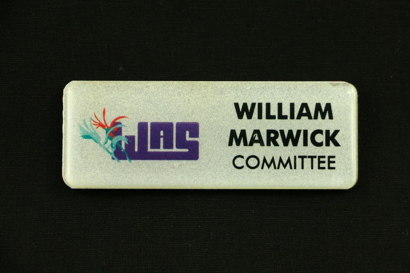 Rectangular, silver coloured plastic name badge with rounded corners. The face of the badge is glittery silver overlaid with thick clear plastic. On the left hand side is a logo featuring an purple acronym that reads 'WAS'. To the left of the acronym are two stylised kangaroo paws, one red and dark green in the background, and one light green and dark green in the foreground. On the right hand side of the badge is black text which reads 'WILLIAM MARWICK COMMITTEE'.