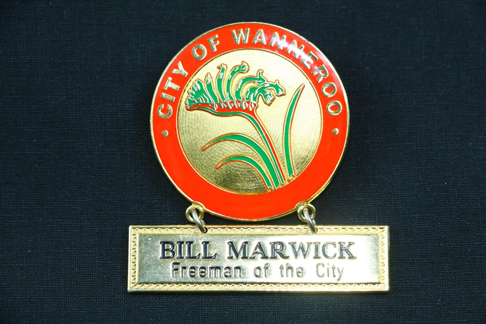 Gold coloured metal badge with enamel circle above a loose rectangular name plate attached by two golden metal chain links. The badge has a red enamel border which reads 'CITY OF WANNEROO' in gold letters. The centre of the circle is a kangaroo paw with three green leaves, a red stem with green flowers, and thin red outlines. The loose nameplate suspended below the circle has a shallow rectangular decorative border embossed with a triangle pattern and reads, 'BILL MARWICK' above 'Freeman of the City'.