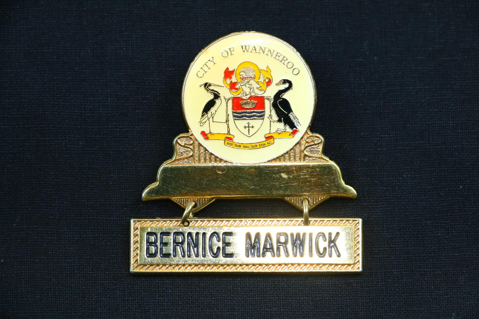 Gold coloured metal badge with a cream coloured enamel circle bearing the City of Wanneroo coat of arms, above a loose rectangular name plate attached by two golden metal chain links. The badge reads 'CITY OF WANNEROO' in small gold lettering above the City's coat of arms. Connecting the bar and the circle are two shallow decorative wedges designed to make the bar resemble a ribbon curling backwards. The loose name plate reads 'BERNICE MARWICK'.