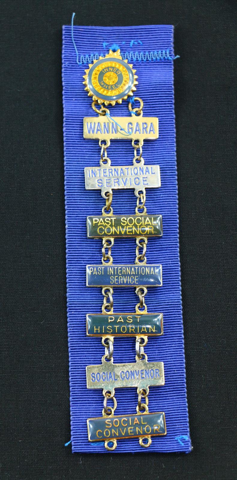 Blue ribbon with multiple badges attached. The first badge is sewn onto the blue ribbon, it is a circle with points around the edge, inside there is a blue outer ring, a gold inner ring, a smaller blue inner ring which has 'INNER WHEEL' written in gold and a small gold centre. There are 7 rectangle badges attached to each other.