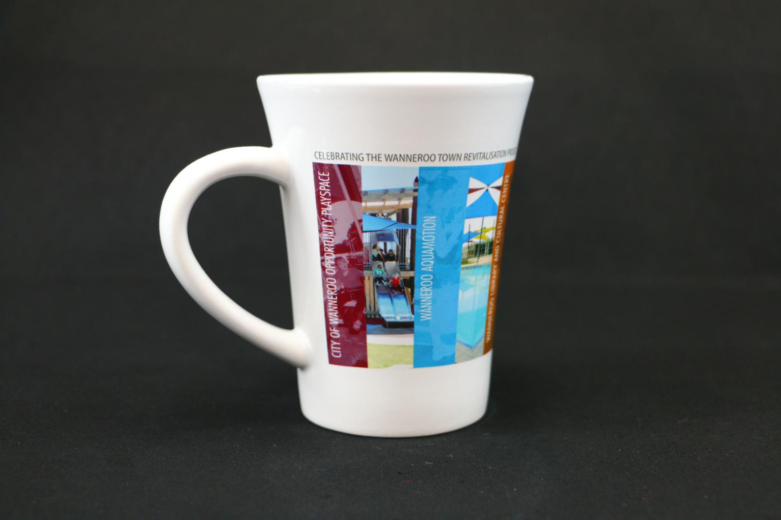 White, ceramic, City of Wanneroo mug with multiple photographic outlines of council locations printed onto the surface around the mug. The top reads 'CELEBRATING THE WANNEROO TOWN REVITALISATION PROJECT'. The first image depicts 'CITY OF WANNEROO OPPORTUNITY PLAYSPACE' and the second image depicts 'WANNEROO AQUAMOTION' .