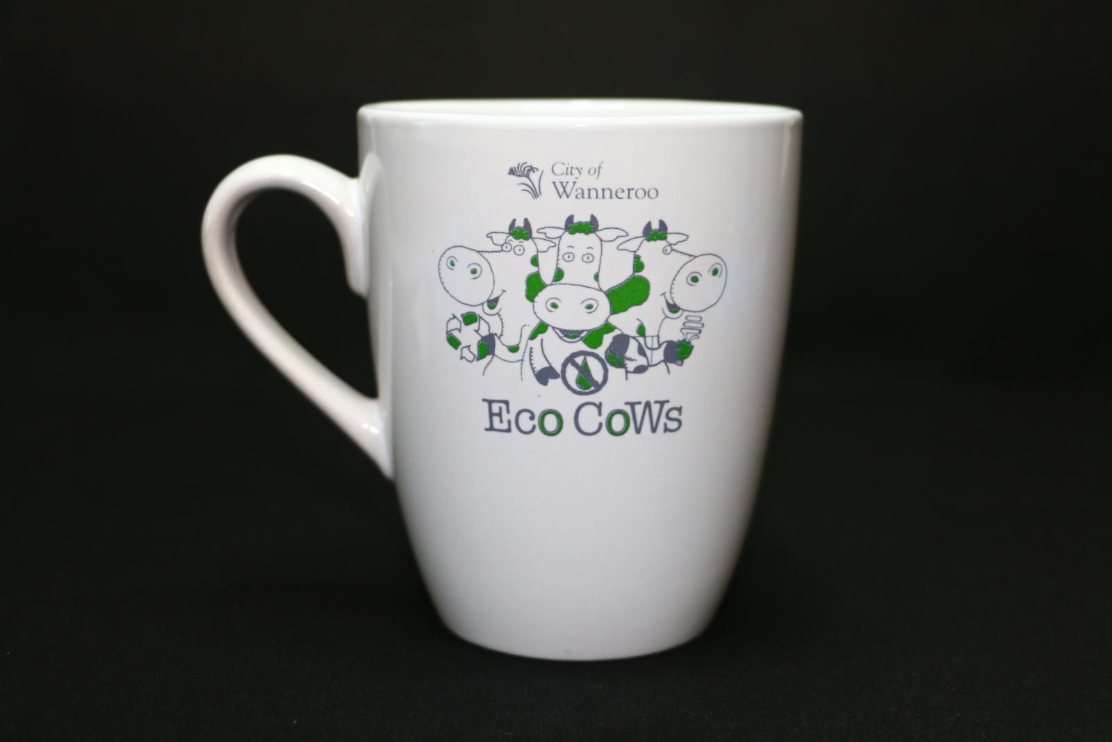 White, ceramic, City of Wanneroo Eco Cows mug. On the front of the mug near the top in small grey letters is 'City of/Wanneroo'. A small grey kangaroo paw flower motif is on the left. Underneath is a motif of three outlines of cartoon cows in grey and green, one looking right, one left, and one towards the middle. Underneath the cows in thick grey and green letters is 'ECO COWS'. This complete motif is replicated exactly on the opposite side of the mug.