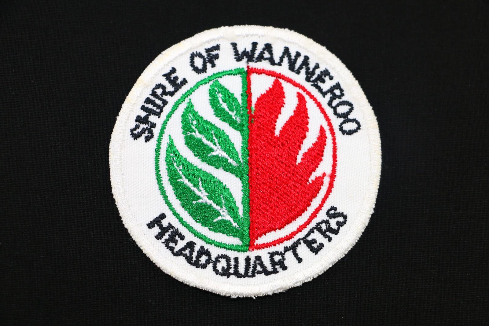 A round Shire of Wanneroo cloth badge. The badge is white and has the words 'SHIRE OF WANNEROO' embroidered across the top and the word 'HEADQUARTERS' at the bottom in black thread. Within these words, there is another circle which has three leaves in green thread with white veins on the left and flames in red thread on the right.  