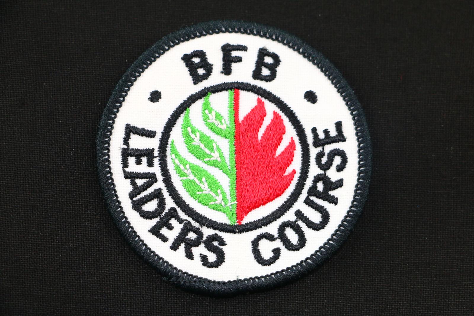 A circular white fabric BFB cloth badge. In a circular design around the edge of the badge the words  'BFB LEADERS COURSE' is embroidered in black thread. Within the circular words is a circular logo which has three leaves in green thread with white veins on the right and flames in red thread on the left side.