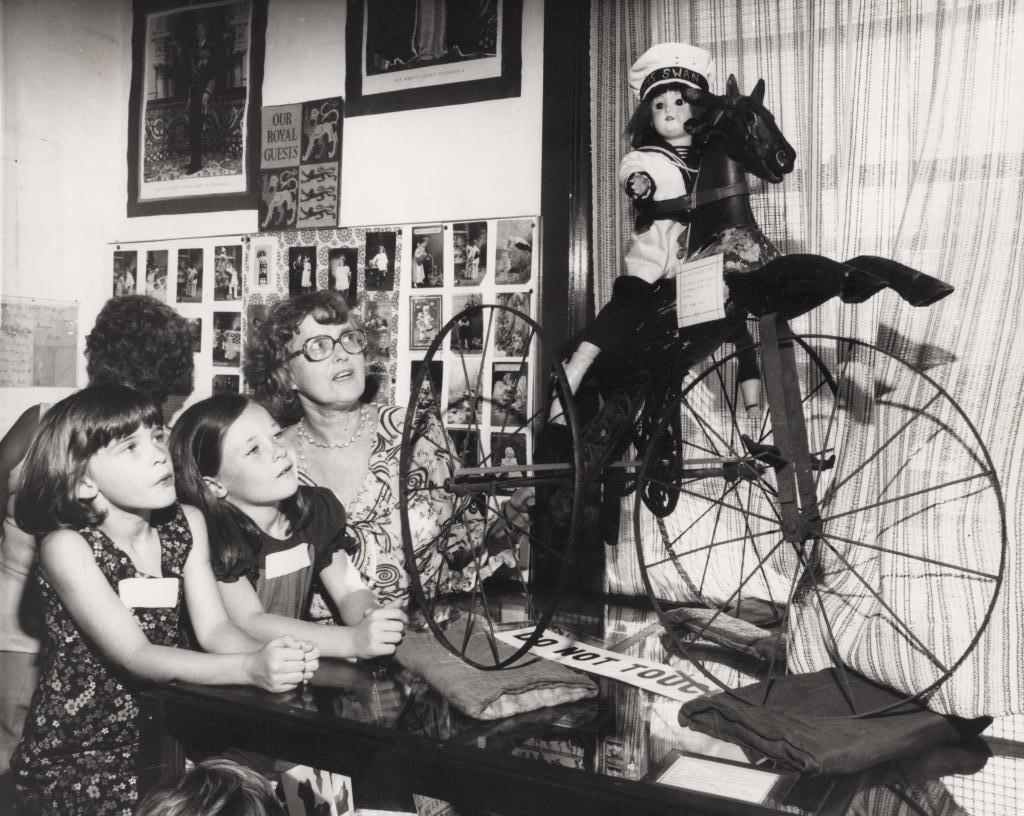 Black and white photograph of a woman wearing glasses and two children who both look at a toy bike with a horse