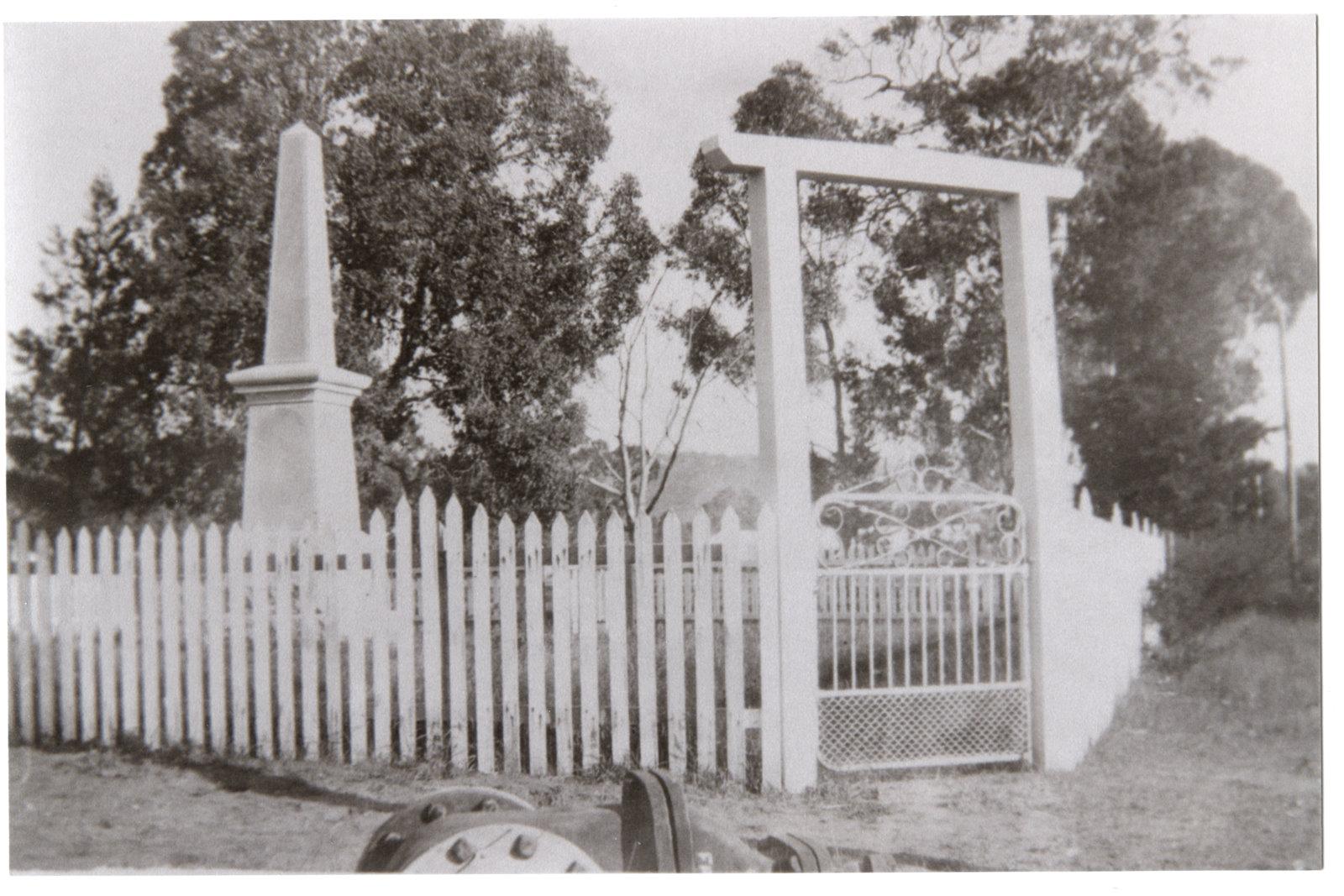 Black and White Photograph of a obelisk shaped War memorial inside a white wooden fence with a rectangle shaped wooded gateway