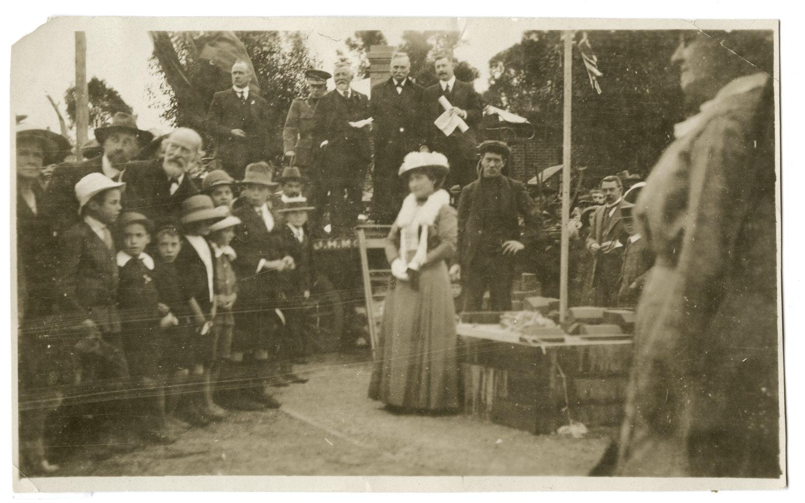 Black and white photograph of a group of people looking at a woman in a dress and hat stranding next to the base of a soon to be built war memorial. Man in workers cloths standing behind her.