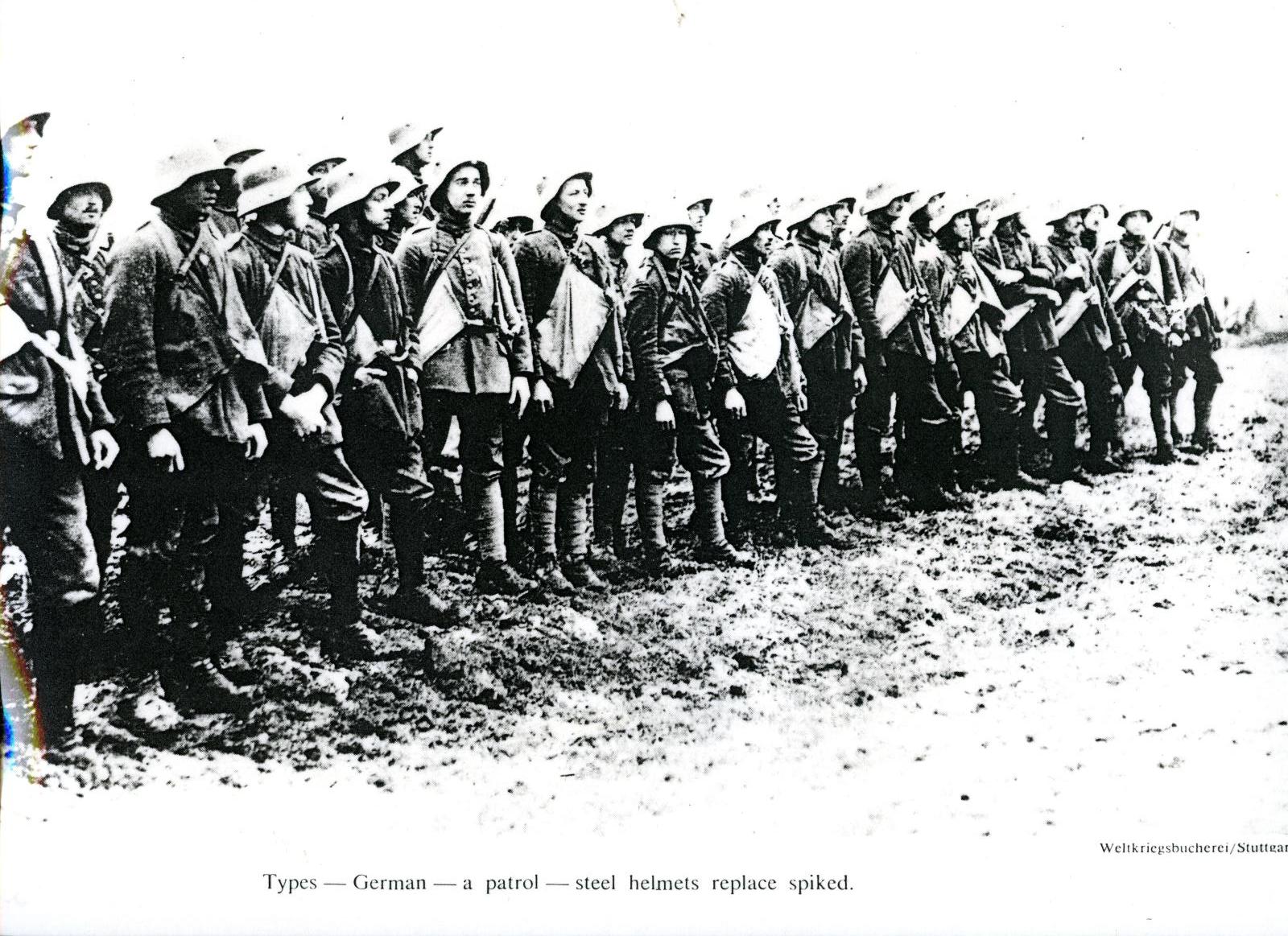 This is a black and white photograph showing a group of German soldiers from World War I, lined up in formation. They are wearing military uniforms and steel helmets, which have replaced the older spiked helmets (Pickelhaube) traditionally worn by German troops. The soldiers appear to be standing in a muddy or rough terrain, and they are carrying their gear, likely preparing for patrol or inspection. The caption at the bottom reads: "Types — German — a patrol — steel helmets replace spiked."  This image cap