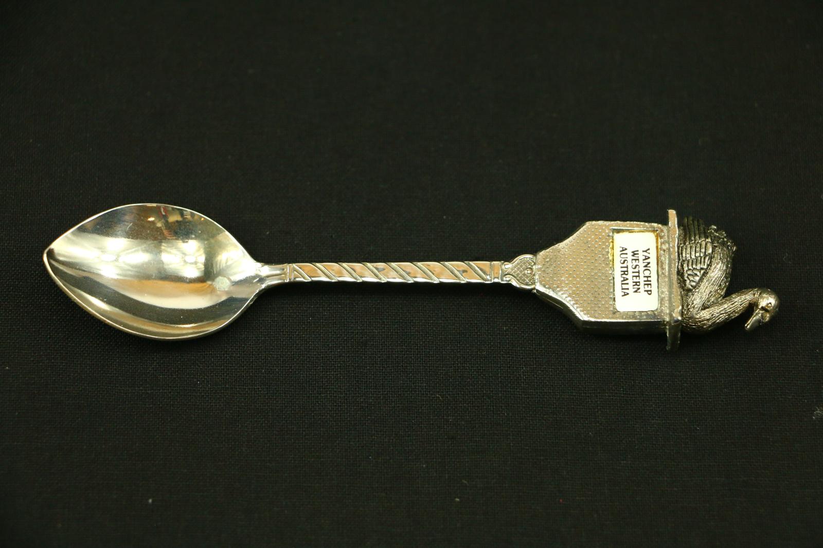 A metal teaspoon with a spiral pattern on the stem which rounds at top with a criss-cross pattern and a dot in the middle. The next section curves out to a square with impressed dot pattern and then a white enamelled oblong shape and the words printed in black 'YANCHEP/WESTERN/AUSTRALIA'. An oblong length ridge of metal sits out over square with a cast finial of a swan facing towards the right.  